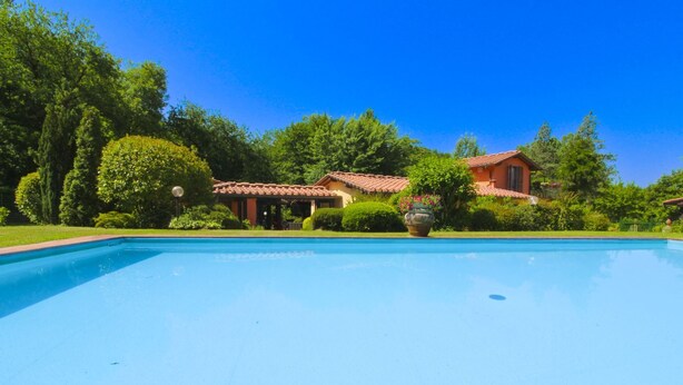A swimming pool with a clear blue sky, surrounded by lush greenery and a house with a red-tiled roof.