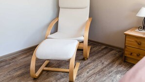 A room with a wooden rocking chair, a small wooden nightstand with a lamp, and a wooden floor.