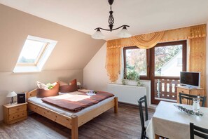 A bedroom with a wooden bed, a desk, a chair, a radiator, a window with curtains, and a TV.