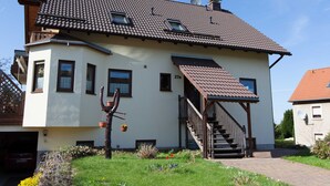 A two-story house with a tiled roof, a staircase leading to the upper floor, and a small garden area with flowers and plants.