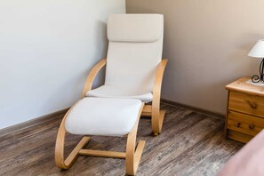 A room with a wooden rocking chair, a small wooden nightstand with a lamp, and a wooden floor.