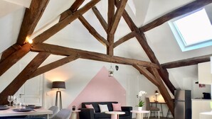 Un habitación estilo loft con vigas de madera a la vista, un comedor con mesa y sillas, un sofá, y una zona de cocina con refrigerador.