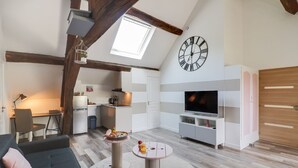 Un salón moderno con un sofá, una televisión y una mesa de comedor con comida. Se aprecia una cocina al fondo con un refrigerador y una ventana.