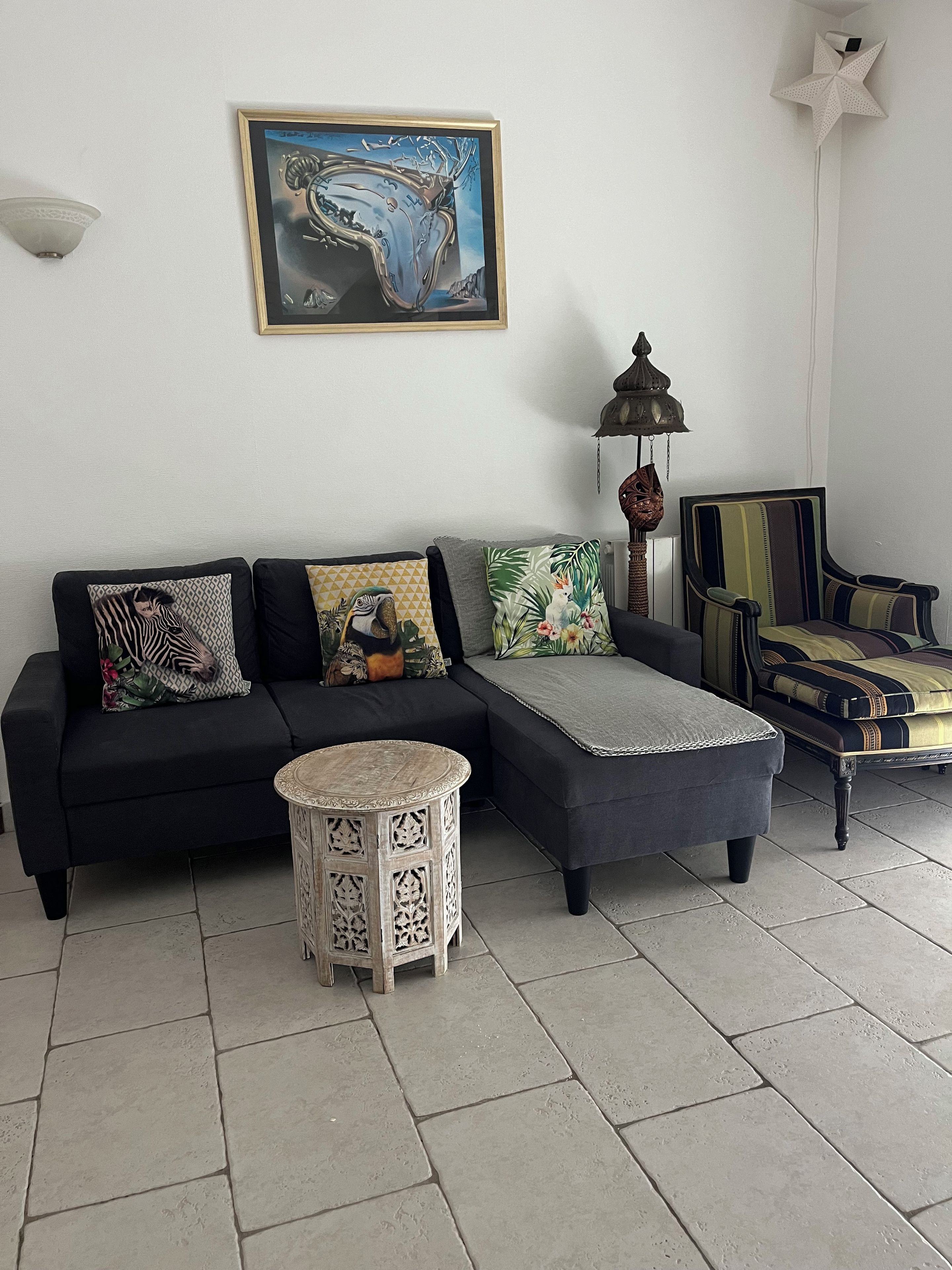 A living room with a black sofa, a round side table, a patterned throw pillow, a star-shaped wall decoration, and a lamp.
