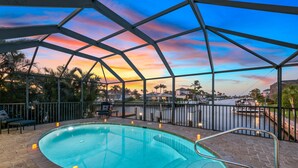 A swimming pool with a view of a sunset over a body of water, surrounded by palm trees and a covered area with seating. - Waterview 3 ensuite home with pool 
HGTV chose as ideal vacation home! (Marco Island)