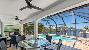 An outdoor patio with a glass table, wicker chairs, a ceiling fan, and a view of a swimming pool and marina. - Waterview 3 ensuite home with pool 
HGTV chose as ideal vacation home! (Marco Island)