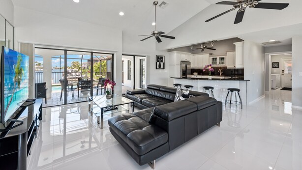 A spacious living area with a black leather sofa, a glass coffee table, and a TV. There are two ceiling fans, a kitchen in the background, and large windows offering a view of the outdoors. - Waterview 3 ensuite home with pool 
HGTV chose as ideal vacation home! (Marco Island)