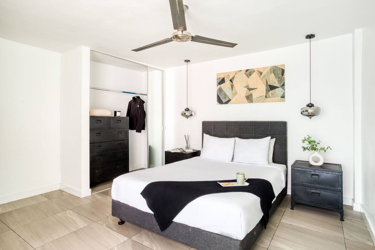 Un dormitorio moderno con una cama grande, un ventilador de techo, una mesita de noche con una planta y una obra de arte en la pared.