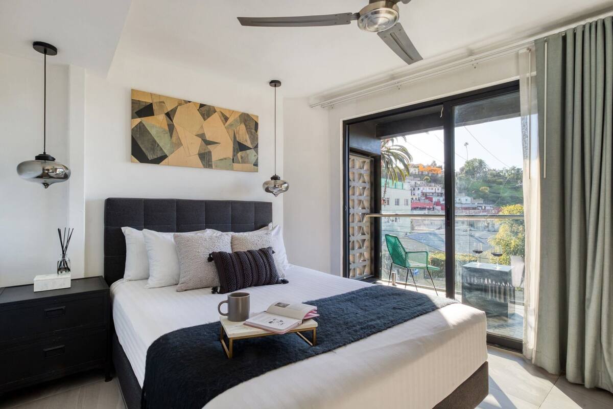 Habitación de hotel moderna con una cama grande, ventilador de techo, vista de la ciudad y balcón con una silla.