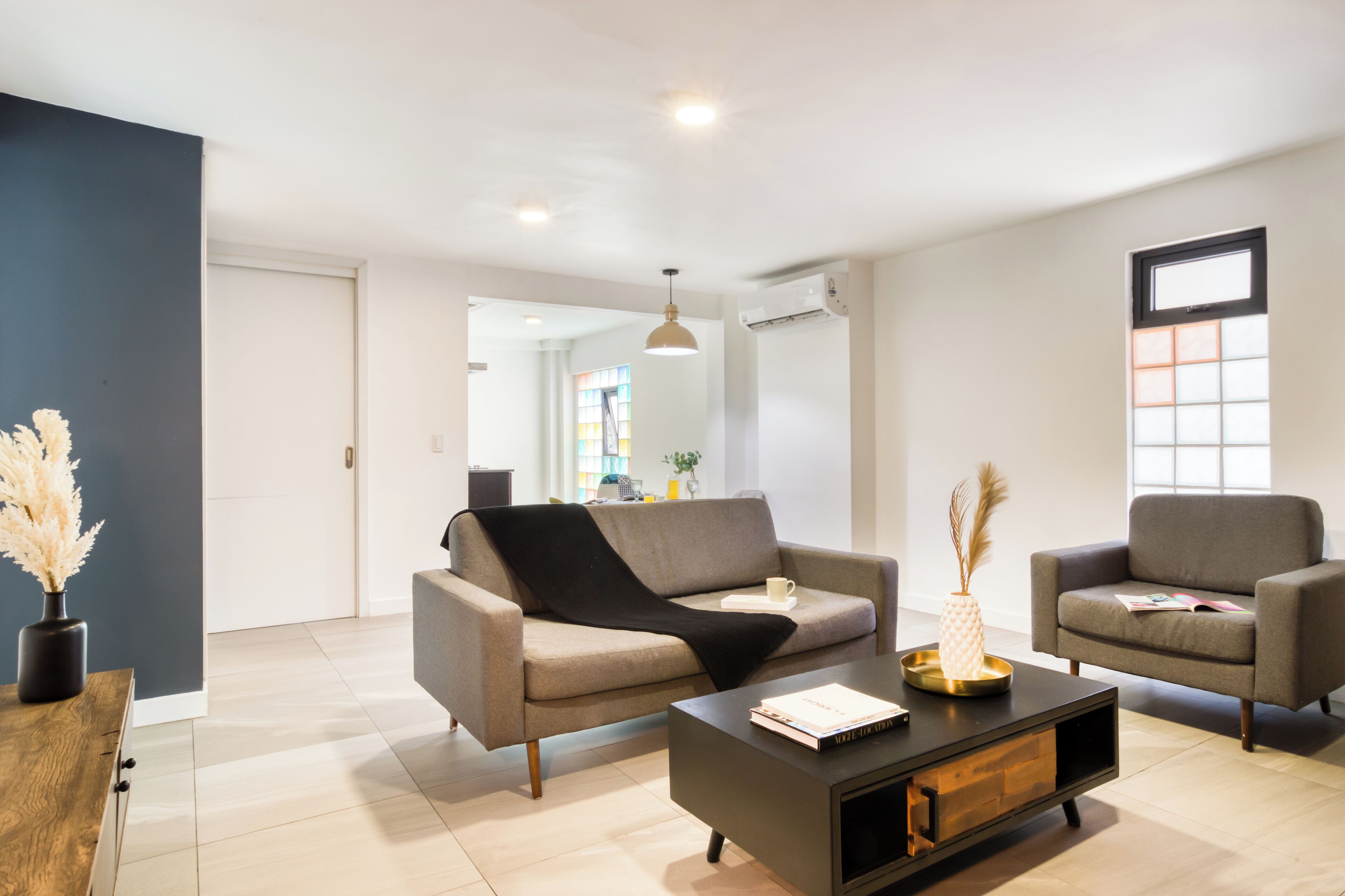 A modern living room with a sofa, armchair, and coffee table. There is a vase with pampas grass, a book, and a remote control on the table.