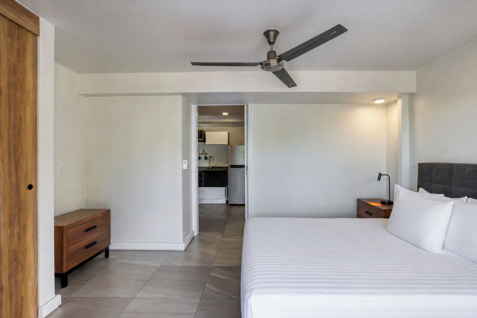 Quarto com cama, mesa de cabeceira, cómoda de madeira, ventoinha no teto e vista para a cozinha.