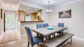 A dining area with a large table, blue chairs, and a bench. There is a kitchen in the background with wooden cabinets and a hanging light fixture.