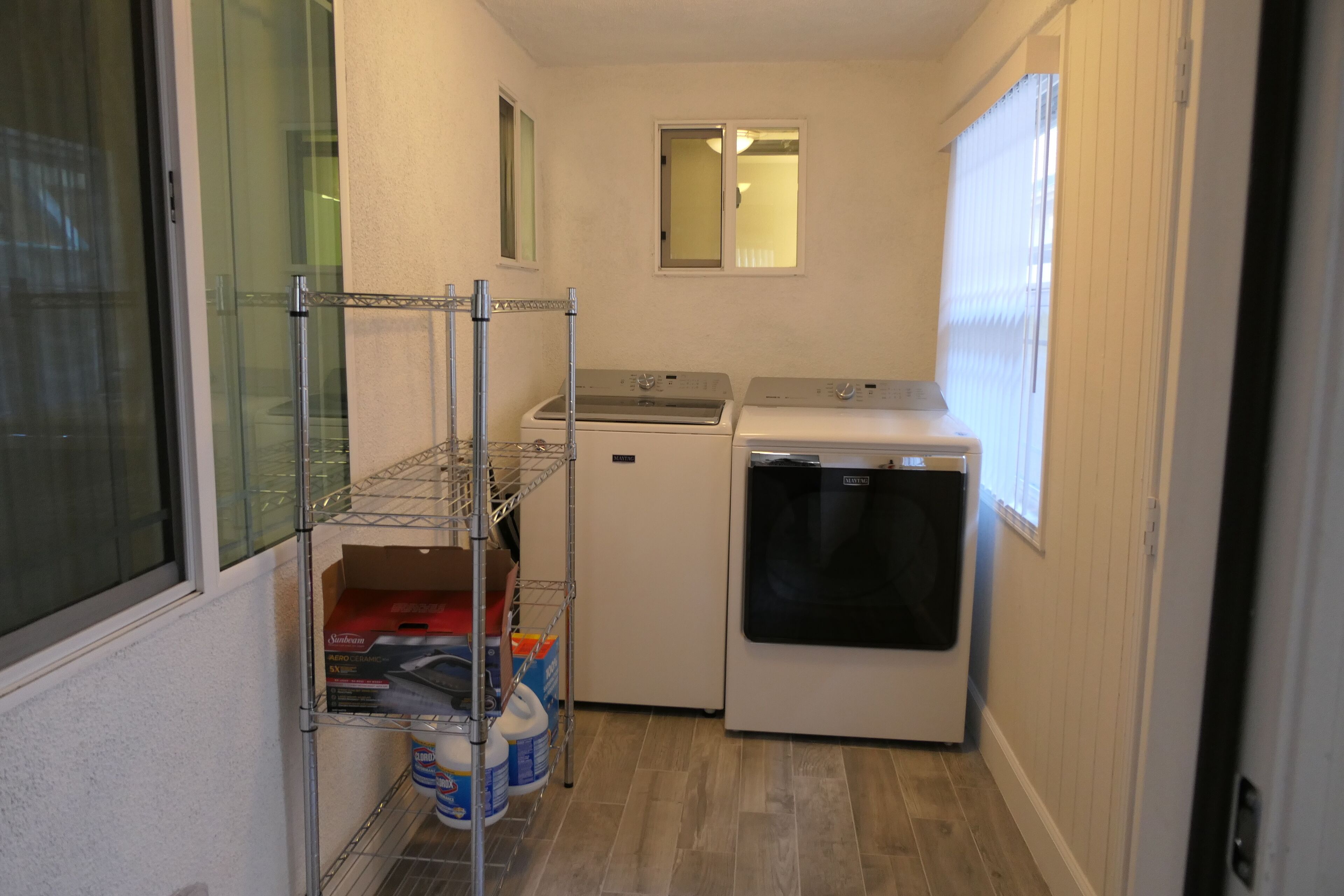 A compact laundry room with a washing machine, dryer, and metal shelving unit with cleaning supplies.