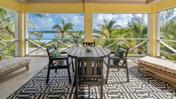 A covered outdoor seating area with a table and chairs, a bench, and a view of the sea and palm trees. - Lo key cottages-The Penthouse (green turtle cay)