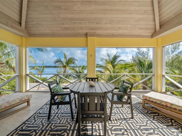 A covered outdoor seating area with a table and chairs, a bench, and a view of the sea and palm trees.