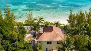 A beachfront property with a single-story building, surrounded by lush greenery and palm trees, overlooking clear blue waters.