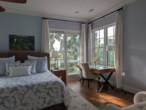 A bedroom with a large bed, a desk with a chair, a painting on the wall, and large windows with curtains.