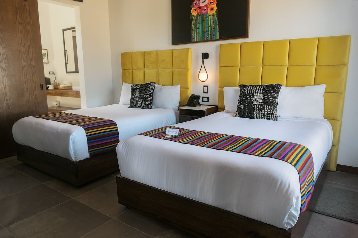 Two double beds with white linens and colorful striped bedspreads, a yellow headboard, and a bedside table with a lamp.