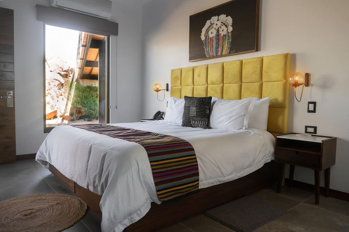 A bedroom with a bed, a yellow headboard, bedside lamps, a painting, and a view of greenery outside.