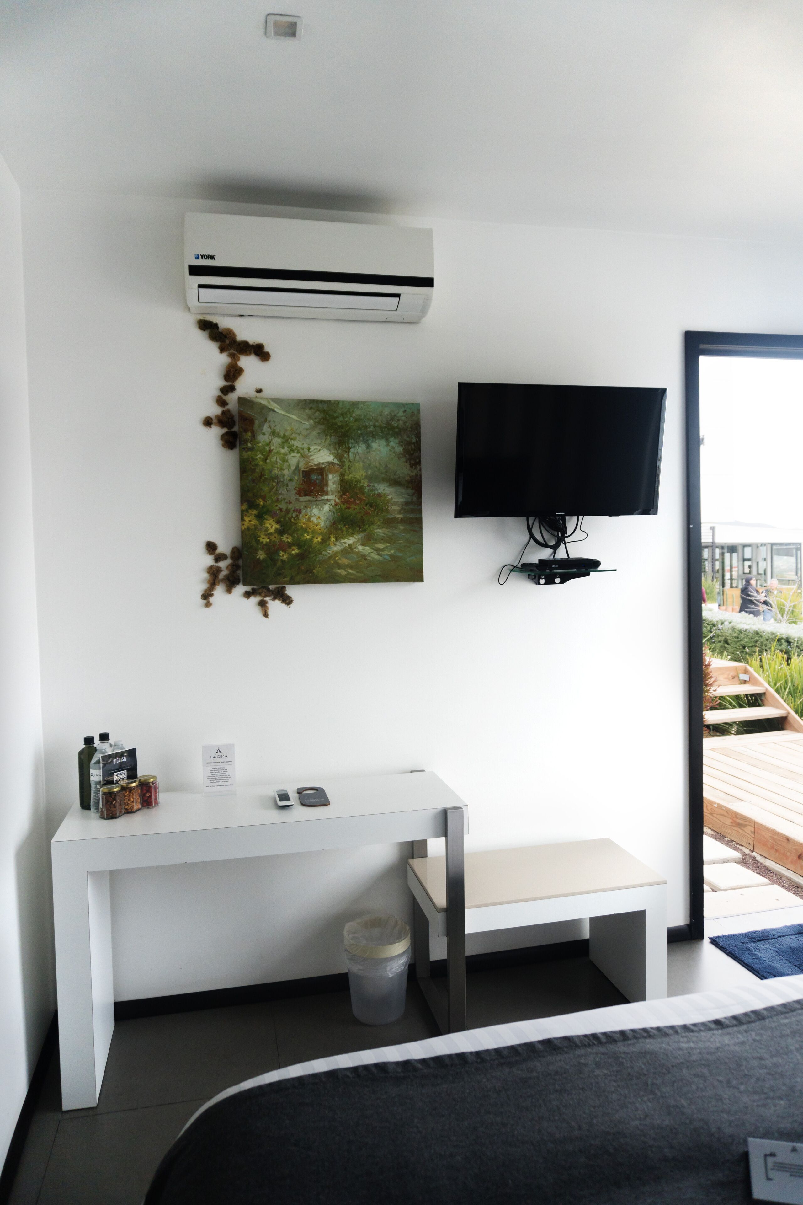 A modern hotel room with a white desk, a mounted flat-screen TV, an air conditioning unit, and a painting on the wall.
