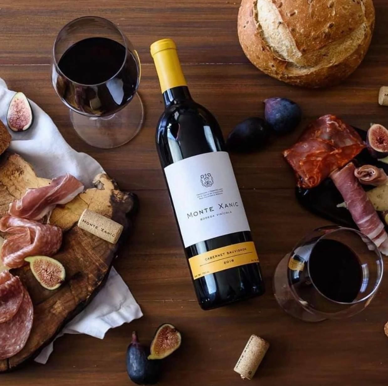 Una botella de vino Monte Xanic, una tabla de madera con diversos embutidos y higos, y dos copas de vino sobre una mesa de madera.