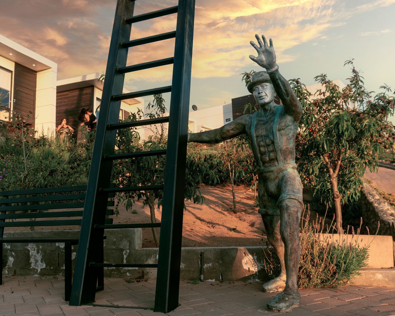 Una estatua de una figura con los brazos extendidos, una escalera y un banco en un área ajardinada con árboles y un edificio al fondo.