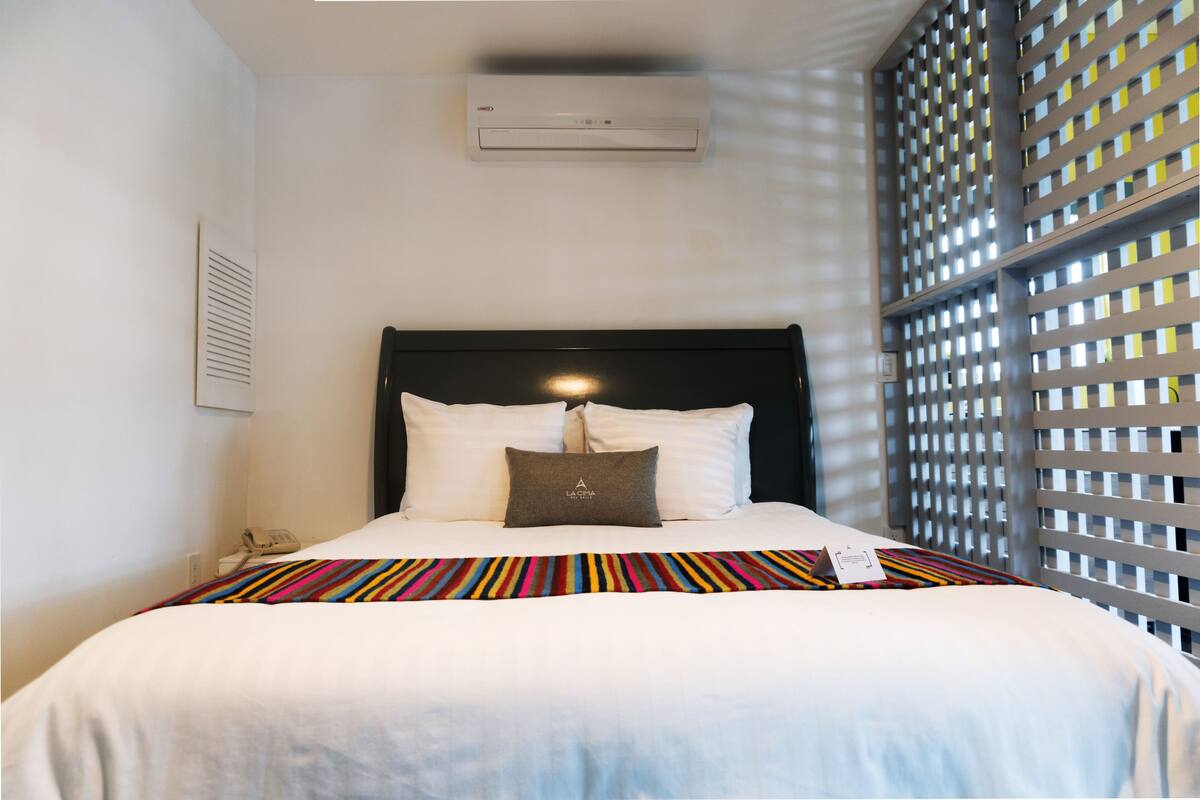 A hotel room with a bed, a white wall, a window with a view, and an air conditioning unit.