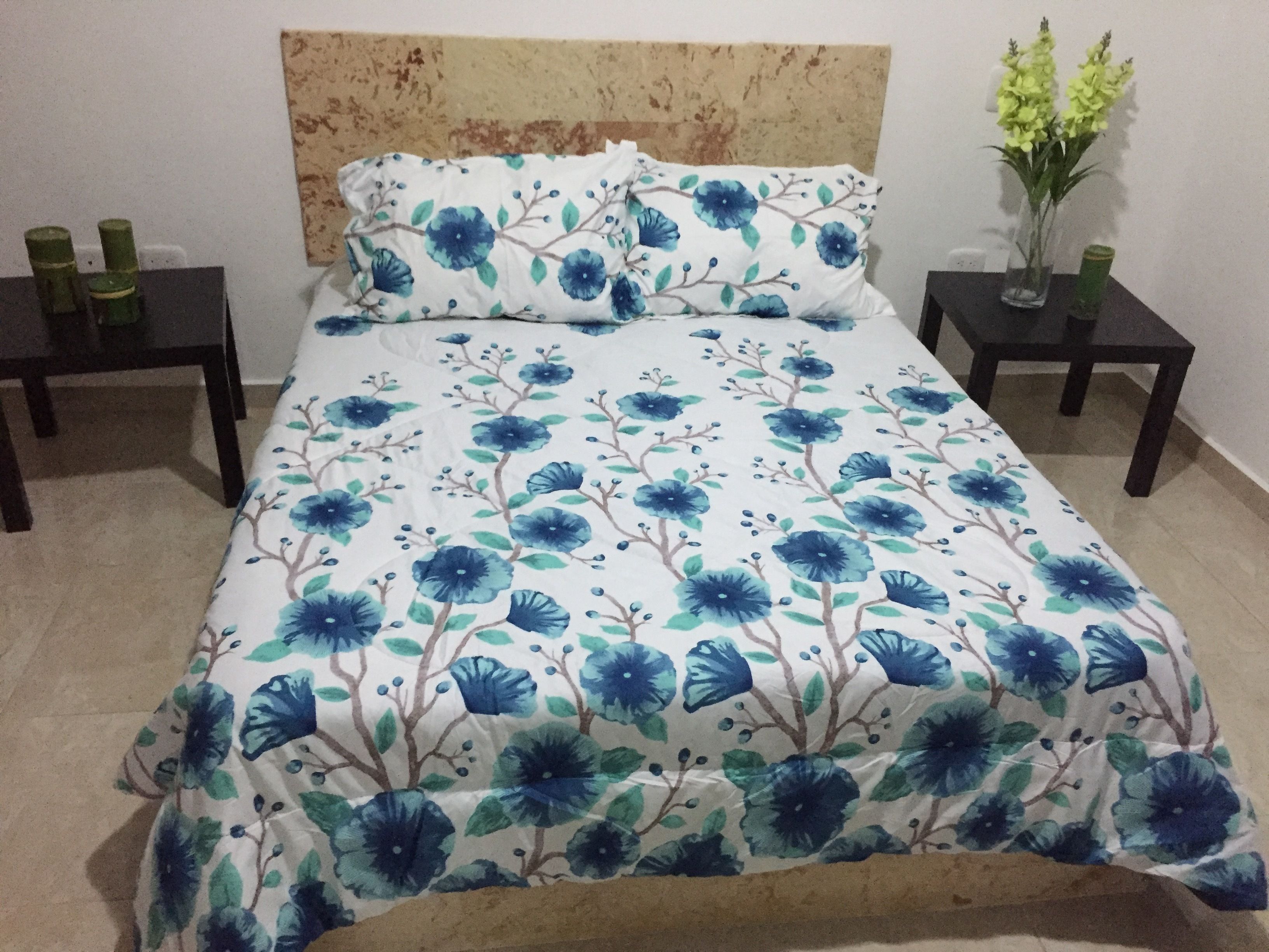 A bed with a floral bedspread, two nightstands with green candles, and a vase with yellow flowers on the right side.