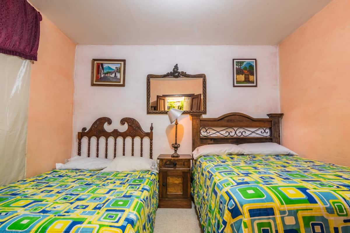 A room with two beds, each with a colorful geometric-patterned bedspread. There is a small bedside table with a lamp between the beds. The walls are painted peach, and there are two framed pictures on the wall above the beds.