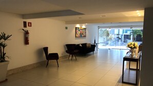 Un hall d’entrée spacieux, avec un canapé noir, un fauteuil, une petite table ornée d’un vase de fleurs et un extincteur fixé au mur.