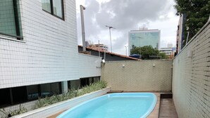 Une piscine rectangulaire dotée d’une terrasse en bois, entourée d’un petit muret en briques et d’un bâtiment aux murs recouverts de carreaux blancs.
