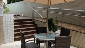 Une terrasse extérieure avec une table ronde et des chaises en osier, un parasol beige et un mur de briques en arrière-plan.
