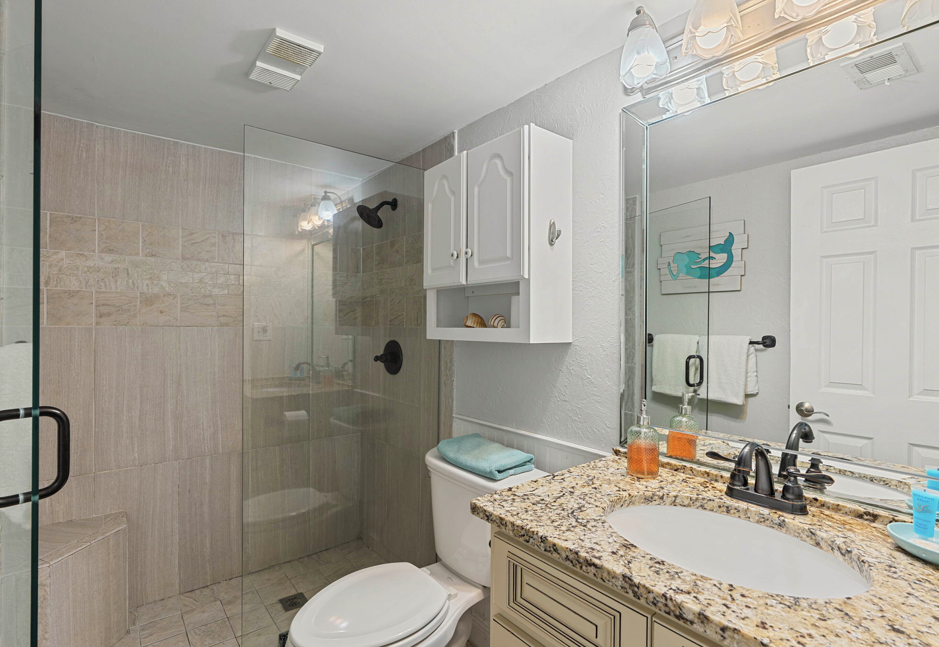 A bathroom with a glass shower enclosure, a white toilet, a granite countertop sink, and white cabinets.