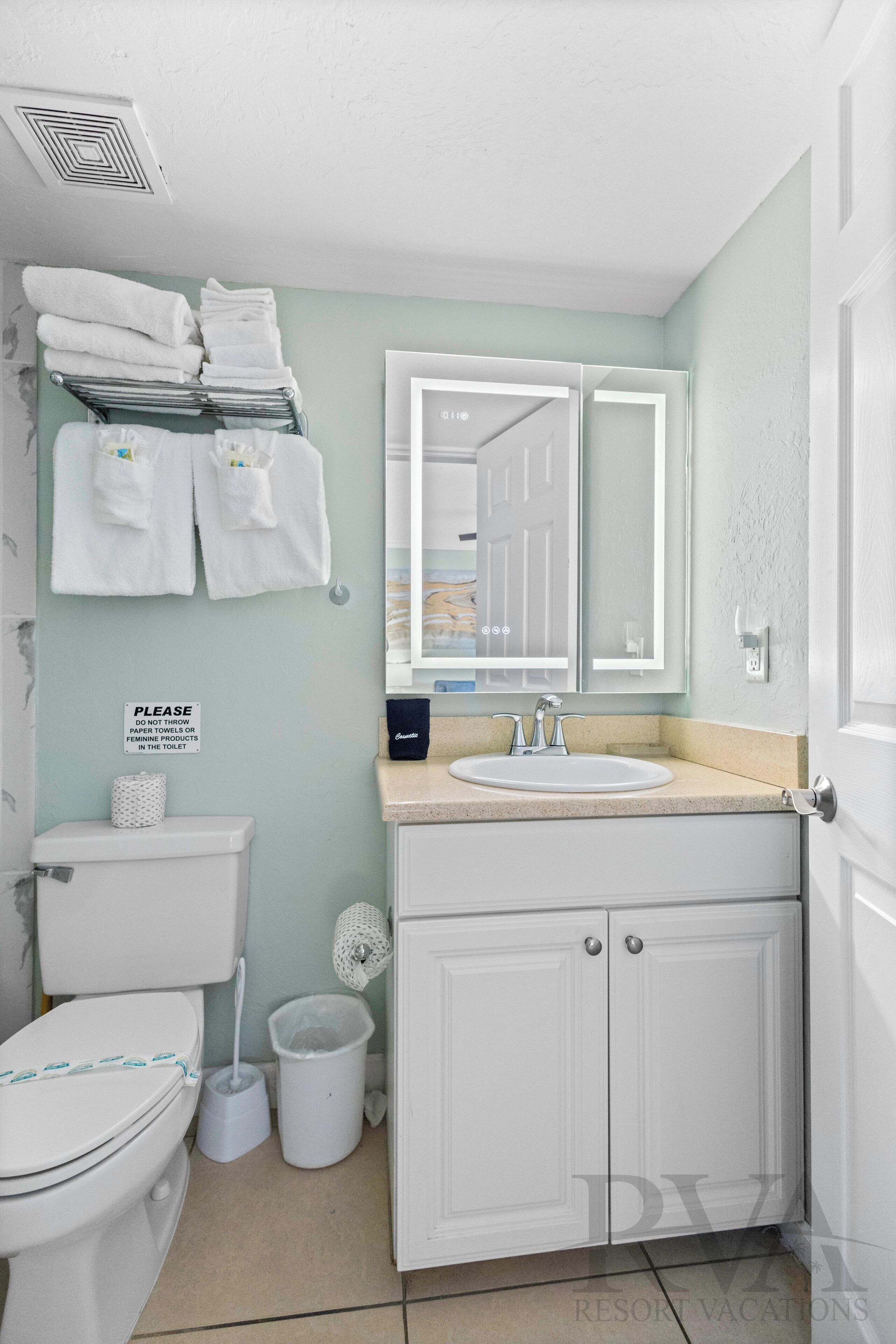 Ein Badezimmer mit Toilette, Waschbecken und Schrank. Handtücher hängen an einem Wäscheständer und ein Schild ist an der Wand angebracht.