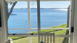 A wooden deck with a table and chairs, a basket with a map and a photo, and a view of a calm body of water with boats. - Beachfront Escape - Tapeka Beach Bach (Tapeka Point (Russell))