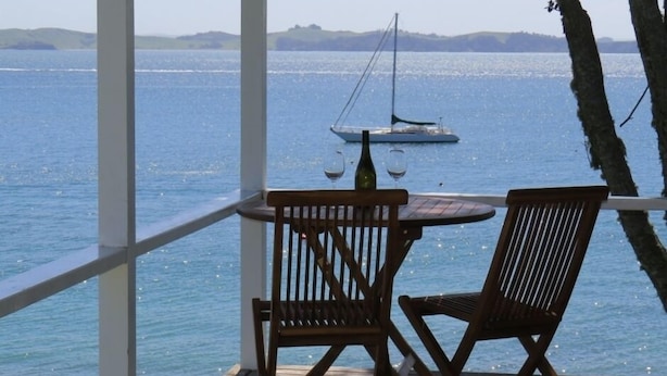 A wooden deck with two chairs and a table, overlooking a calm sea with a sailboat in the distance. - Beachfront Escape - Tapeka Beach Bach (Tapeka Point (Russell))