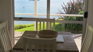A room with a wooden table and chairs, a woven basket, and a view of a lake with boats. - Beachfront Escape - Tapeka Beach Bach (Tapeka Point (Russell))