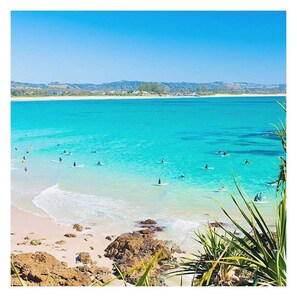 Een strand met helder turquoise water, mensen die zwemmen en surfen, en rotsachtige landtongen.