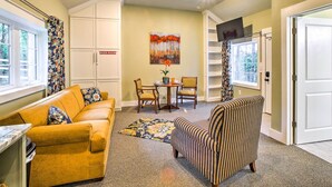 A living room with a sofa, armchair, and a small table with chairs. There is a painting on the wall and a door with a sign. - Steps to Branson Landing: Cozy Downtown Cottage! (Branson)