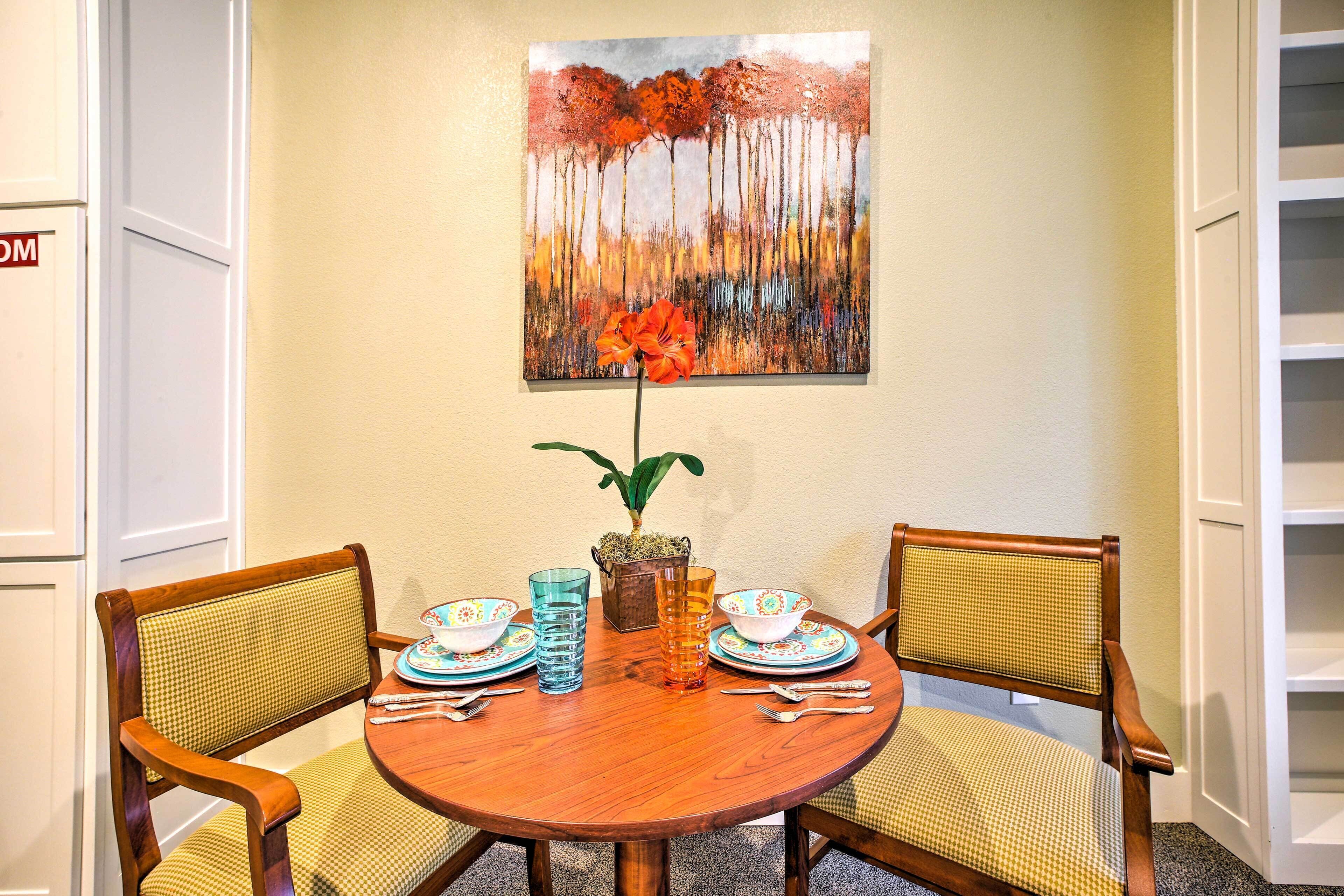 A dining area with a round wooden table, two chairs, a vase with a single flower, and a painting of trees on the wall.