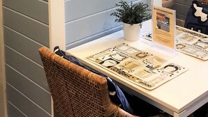 Een kamer met een witte tafel, een rieten stoel, een kleine potplant en een spiegel die een handdoek en een deur reflecteert.