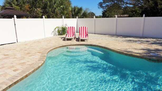 A swimming pool with clear blue water, surrounded by a white fence and two red and white striped lounge chairs.