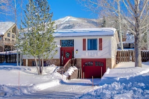 A snow-covered house with a red door and windows, surrounded by trees and a wooden fence.