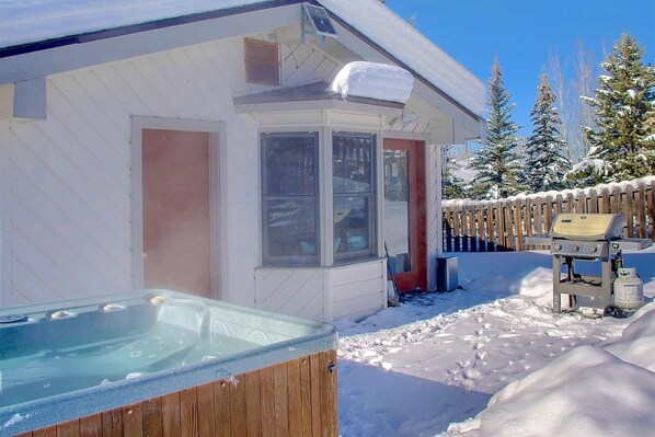Una bañera de hidromasaje de madera al aire libre frente a una casa con una terraza cubierta de nieve y una valla de madera al fondo.