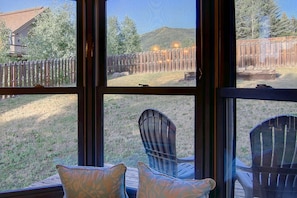 A view from a window with two chairs and a cushion, overlooking a backyard with a wooden fence, a hill, and a house in the background.