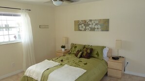 A bedroom with a bed, two bedside tables, a ceiling fan, a window with curtains, and a wall-mounted painting.