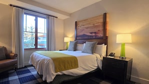 Habitación de hotel con una cama grande, dos lámparas de noche, una silla, un cuadro en la pared y una ventana con cortinas.