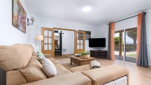 Een moderne woonkamer met een beige bank, een bijzettafeltje en een televisie. Er is een schuifdeur naar een buitenruimte.