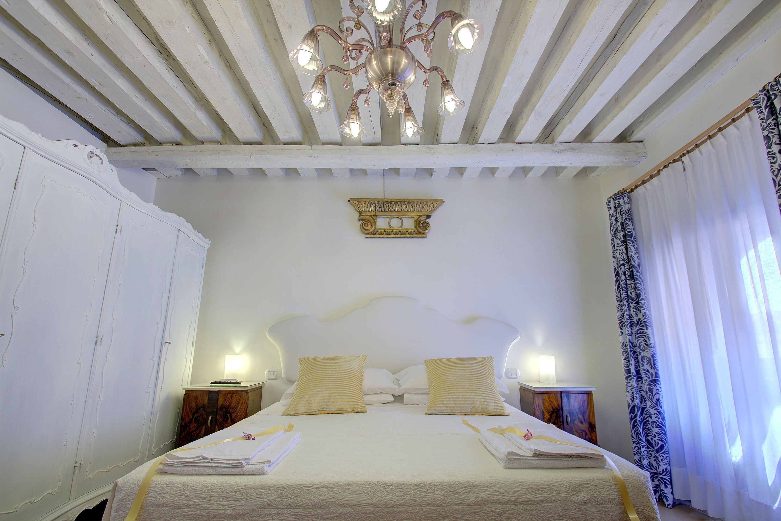 Een keurig opgemaakt bed met witte lakens en twee kussens, een kroonluchter met meerdere lampen, en een houten hoofdbord.