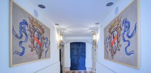 Un couloir au sol à carreaux, orné de deux peintures murales décoratives, et une porte bleue au fond.